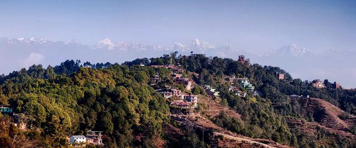 Snow-capped Himalayan peaks floating above haze behind a forested hillside village in morning light