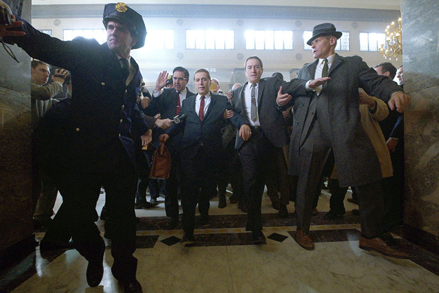 Al Pacino as Jimmy Hoffa strides through a marble lobby with Robert De Niro amid a jostling crowd and police