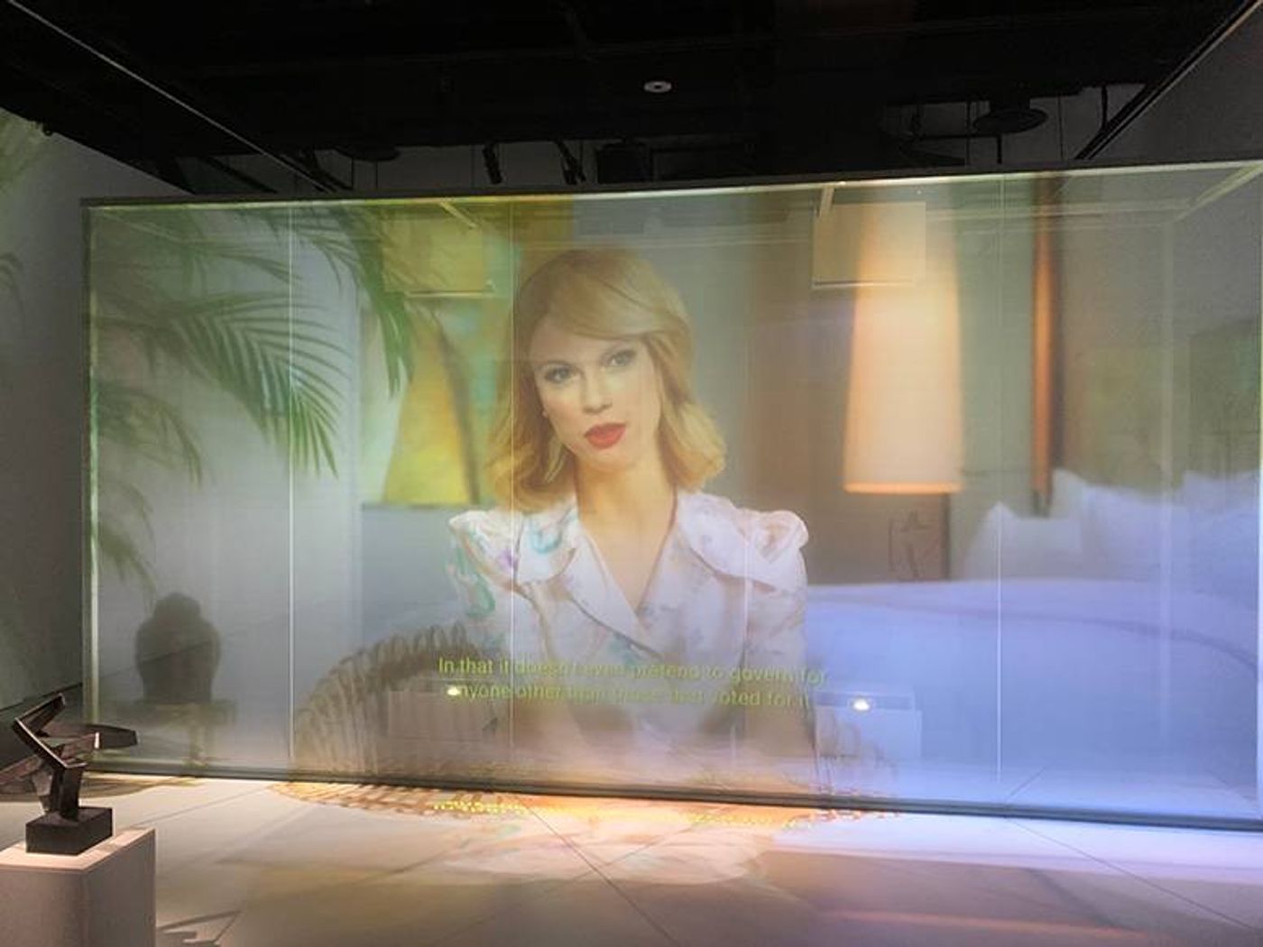 A blonde woman speaks on a translucent video screen in Kulendran Thomas and Kuhlmann’s Being Human