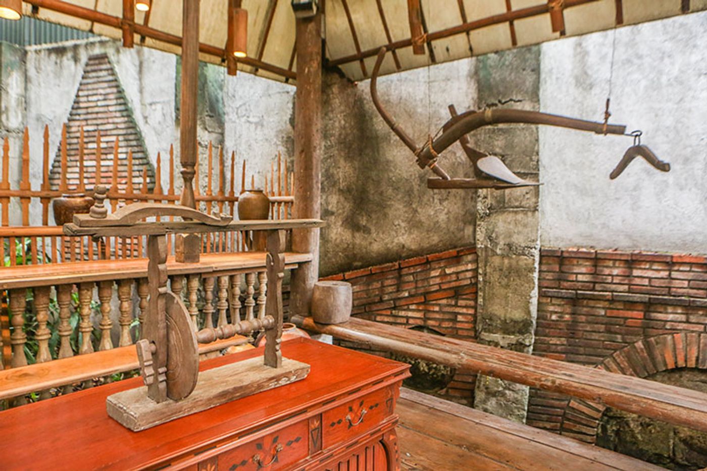 An antique wooden sewing contraption on a red painted cabinet inside the bahay kubo, an old araro plough hanging from the rafters behind it