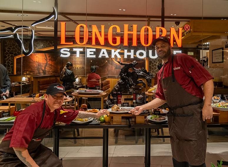 Two smiling crew members in maroon aprons hold out plates beneath the LongHorn Steakhouse sign