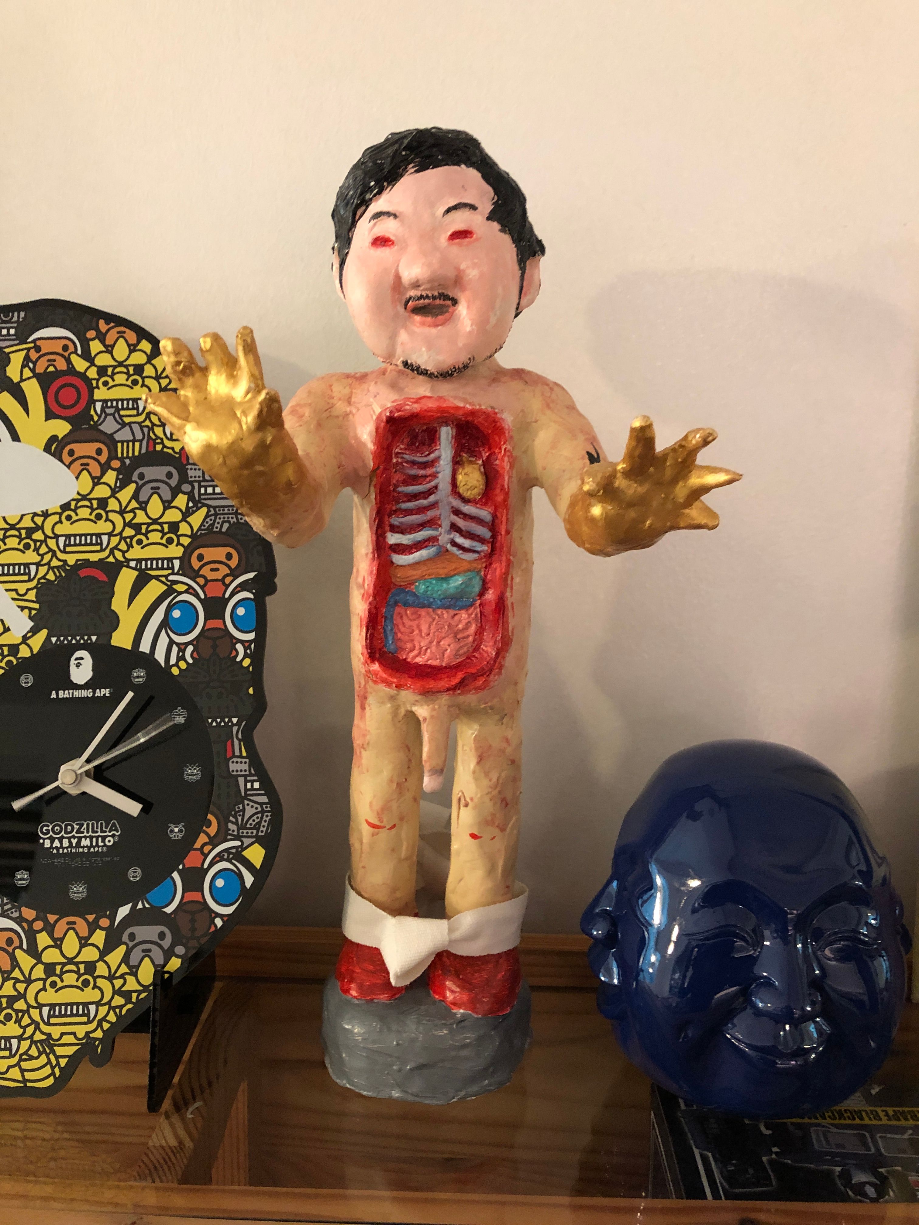 A papier-mâché figure with its chest opened to show ribs and organs and its gilded hands raised, beside a blue ceramic Buddha head and a Bathing Ape Godzilla wall clock