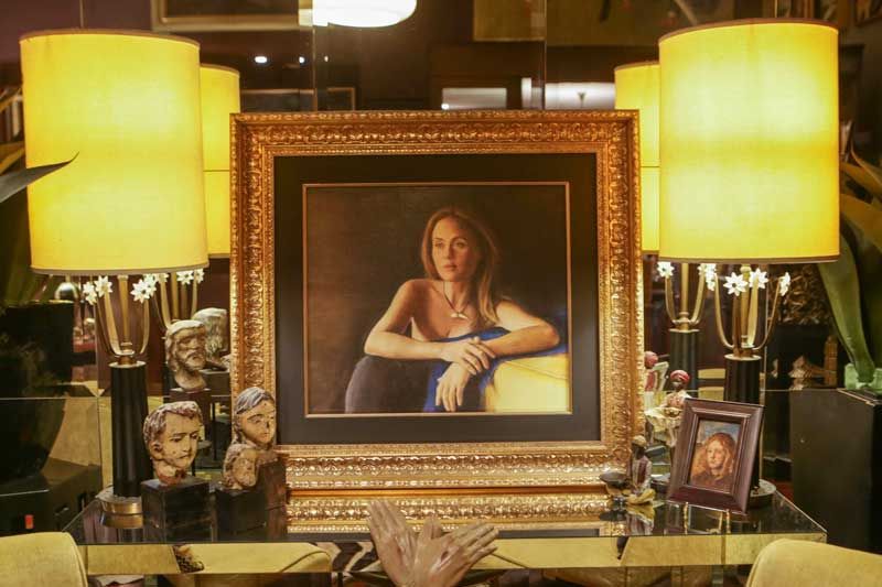 A gold-framed oil portrait of Joanna in a blue dress displayed between two tall lamps among small sculptures