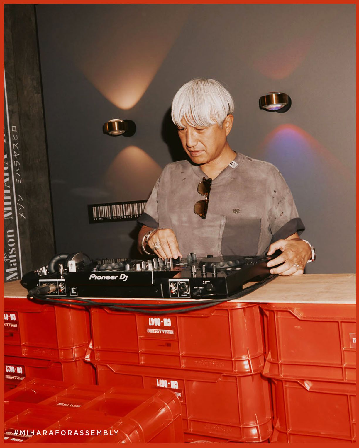 Japanese designer Mihara Yasuhiro in front of a DJ setup, smiling and wearing one of his signature deconstructed sneakers