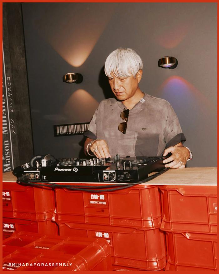 Japanese designer Mihara Yasuhiro in front of a DJ setup, smiling and wearing one of his signature deconstructed sneakers