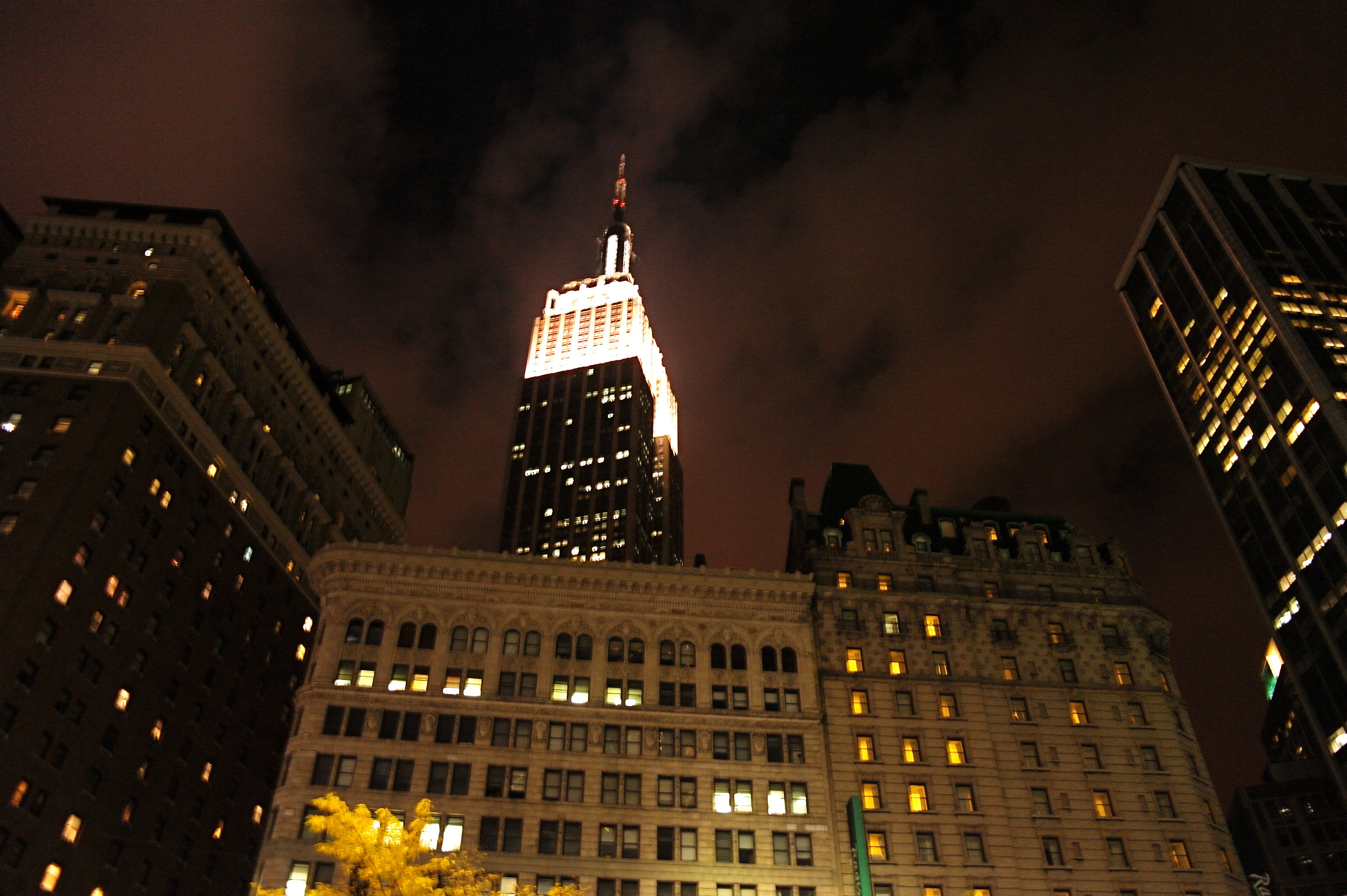 The Empire State Building, its crown floodlit white, shot from the street at night between dark towers and an ornate stone facade full of glowing windows