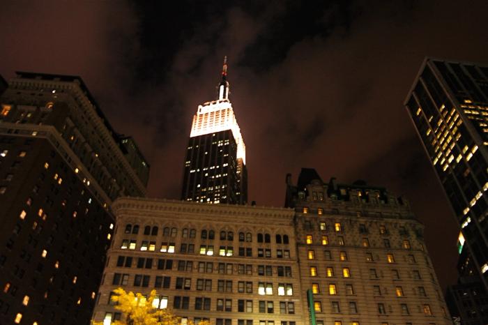 The Empire State Building, its crown floodlit white, shot from the street at night between dark towers and an ornate stone facade full of glowing windows
