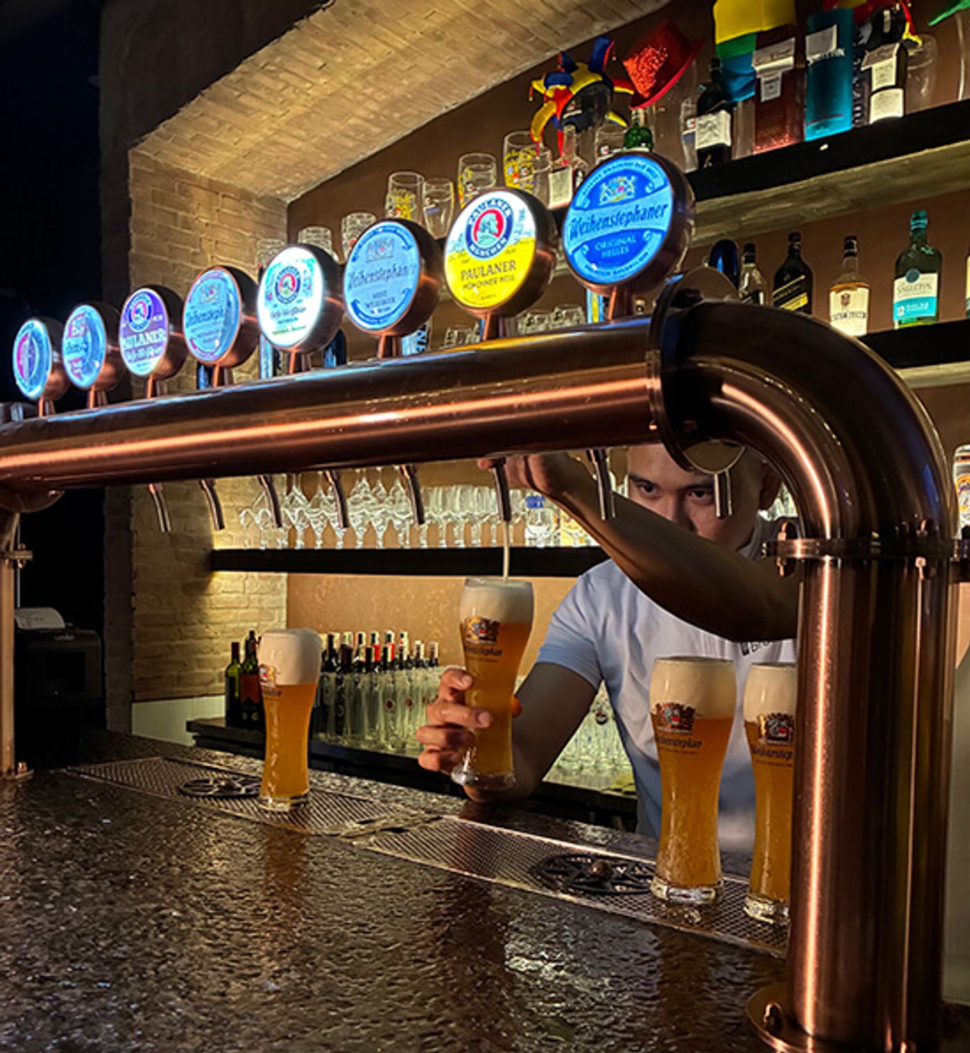 A bartender pours wheat beer from a copper tap tower lined with German beer badges at Brotzeit