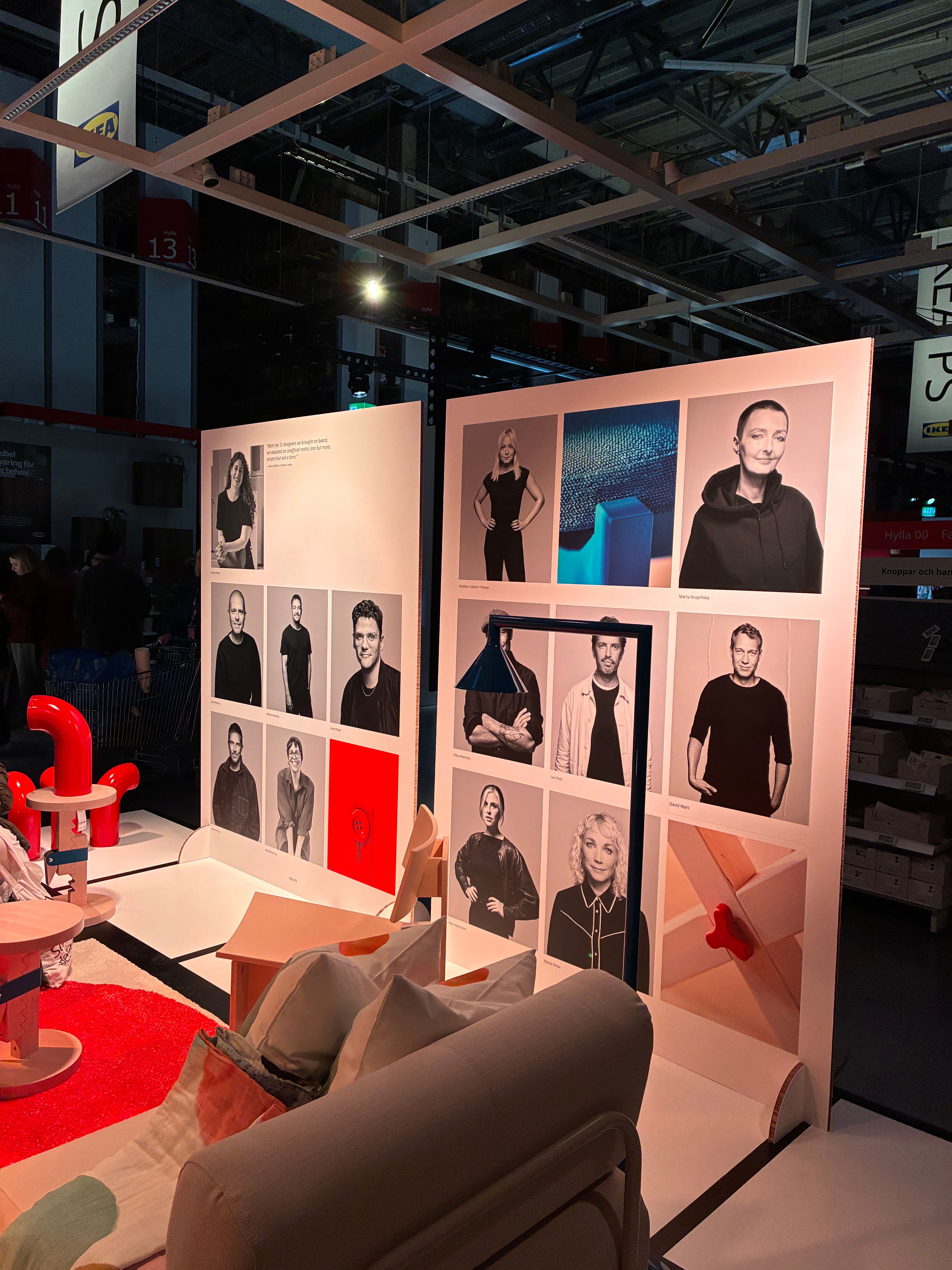Wall of black and white designer portraits at the IKEA PS 2026 exhibition in Älmhult, with red tubular furniture and a pale sofa in front
