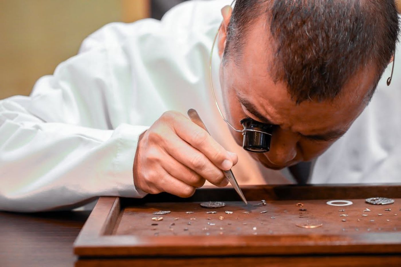 A watchmaker wearing an eye loupe lifts tiny movement parts with tweezers from a wooden tray at Patek Philippe’s Knowledge Series