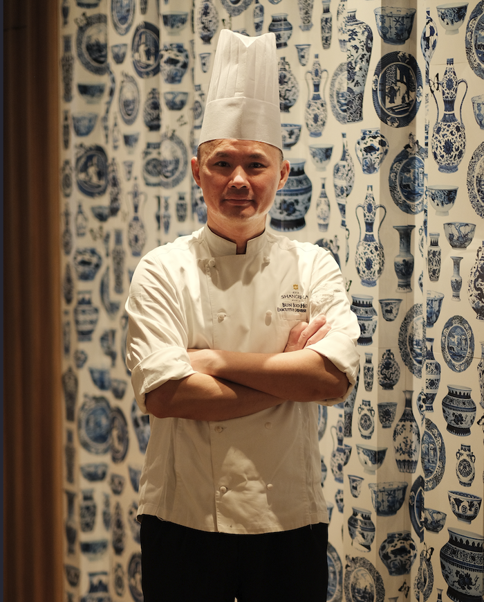 Chef BoonHoe Bun standing with arms crossed in his chef uniform and tall white hat in front of a blue and white porcelain patterned background