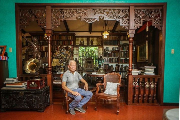 Dr. Jaime Laya sits in the carved narra doorway of his library, bookshelves and santos behind him, a brass gramophone horn and a caned chair beside him