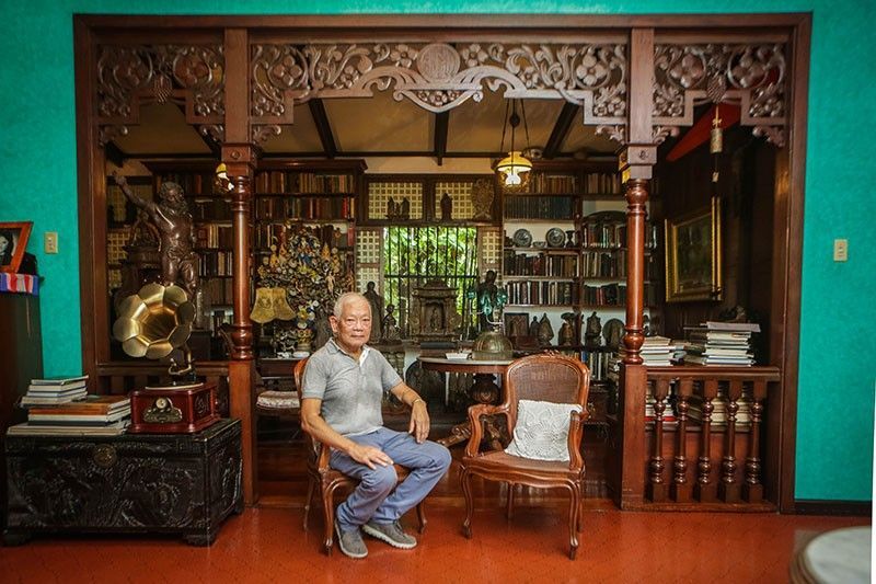 Dr. Jaime Laya sits in the carved narra doorway of his library, bookshelves and santos behind him, a brass gramophone horn and a caned chair beside him