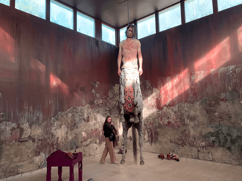 A visitor stands beneath a giant hanged male centaur in the weathered Danish Pavilion