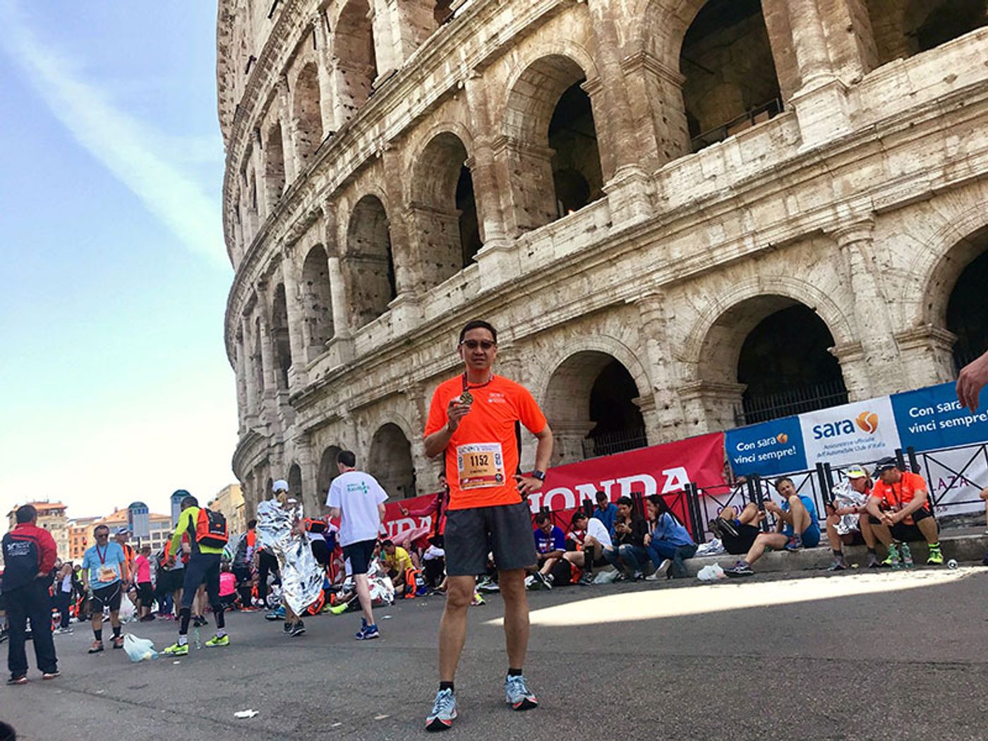 Emerson Yao in an orange race shirt holding up a medal in front of the Colosseum, runners in foil blankets behind him