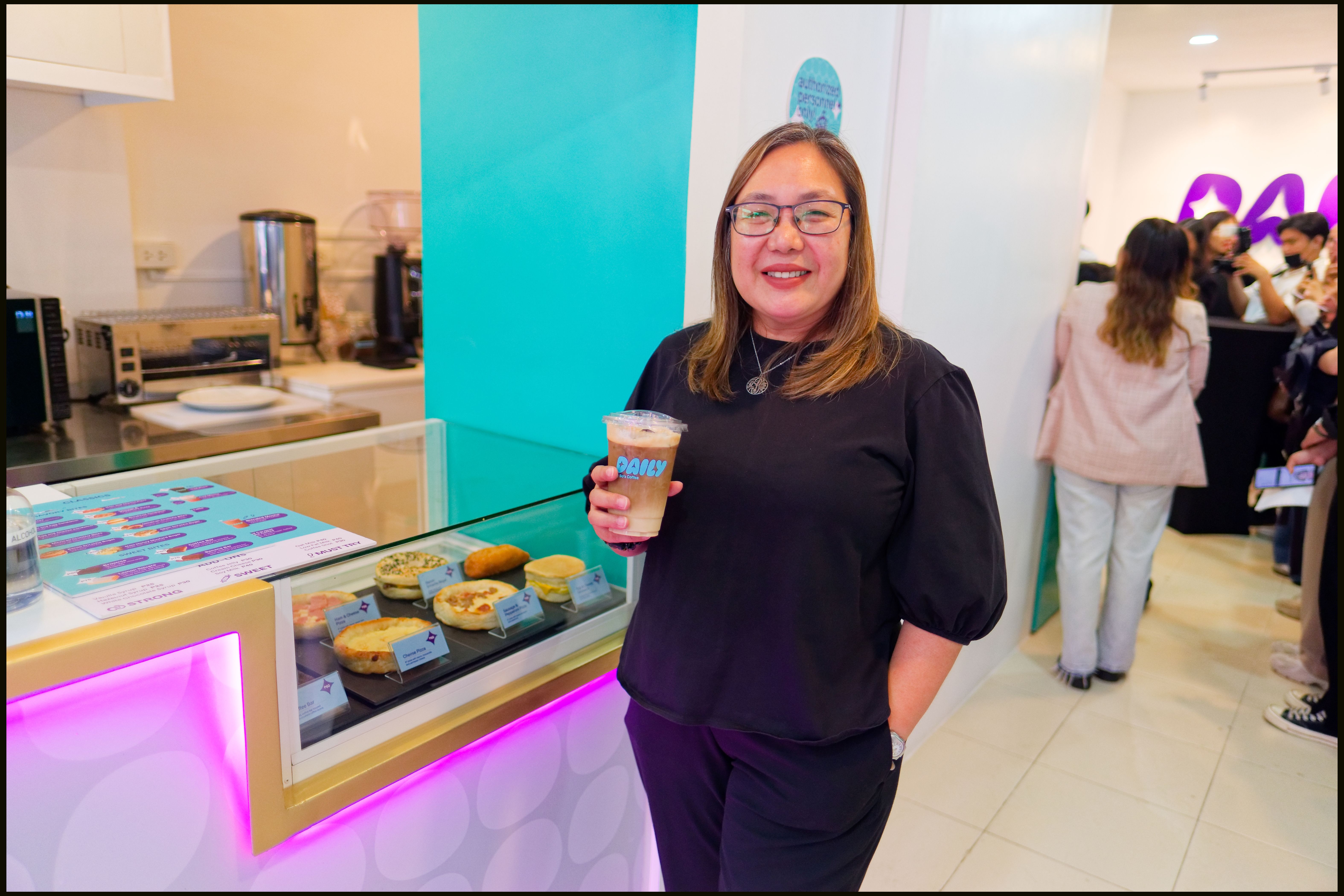 A smiling woman in black holds an iced coffee beside a glass pastry case at the Daily by Bo’s Coffee store