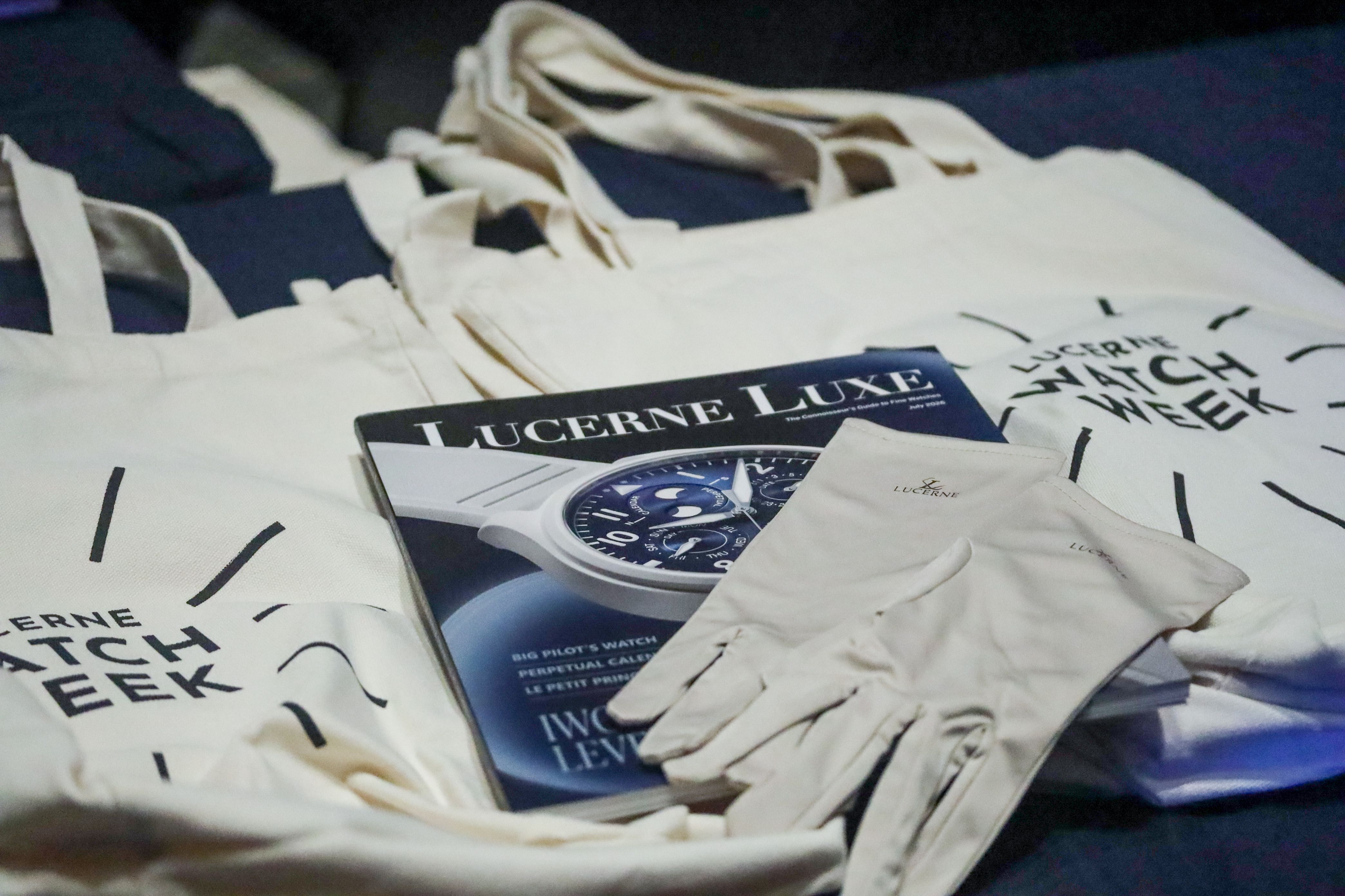 A Lucerne Luxe magazine issue featuring a blue dial watch on the cover, surrounded by white canvas tote bags printed with Lucerne Watch Week and a pair of white watchmaker handling gloves resting on top