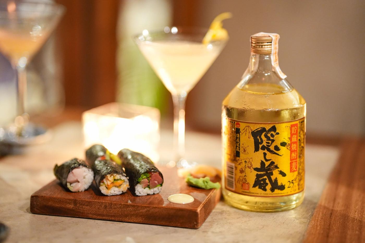 A pale shochu cocktail in a martini glass beside three Sakagura hand rolls on a wooden board and a bottle of Kakushigura barley shochu