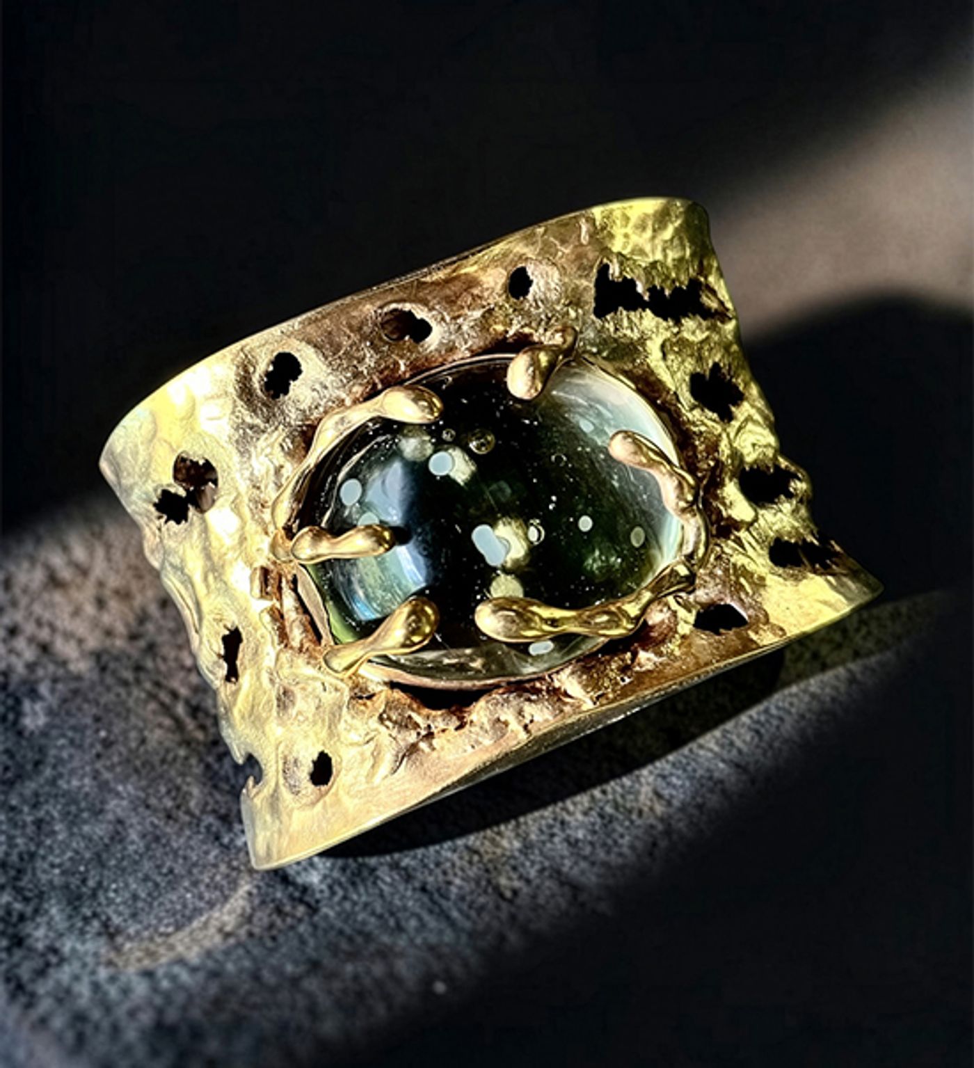 Hand-torched gold cuff by Michelline Syjuco pitted with holes around a dark vintage glass stone