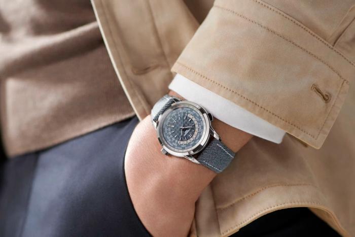 The blue-dialed Patek Philippe World Time watch worn on a wrist below a tan trench-coat cuff