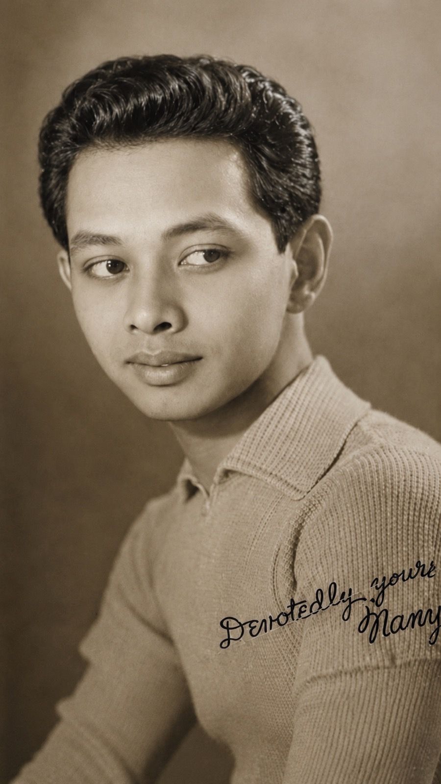 Sepia-toned vintage studio portrait of a young man with wavy hair in a knit collared shirt, a handwritten inscription reading ‘Devotedly yours’ across the shoulder