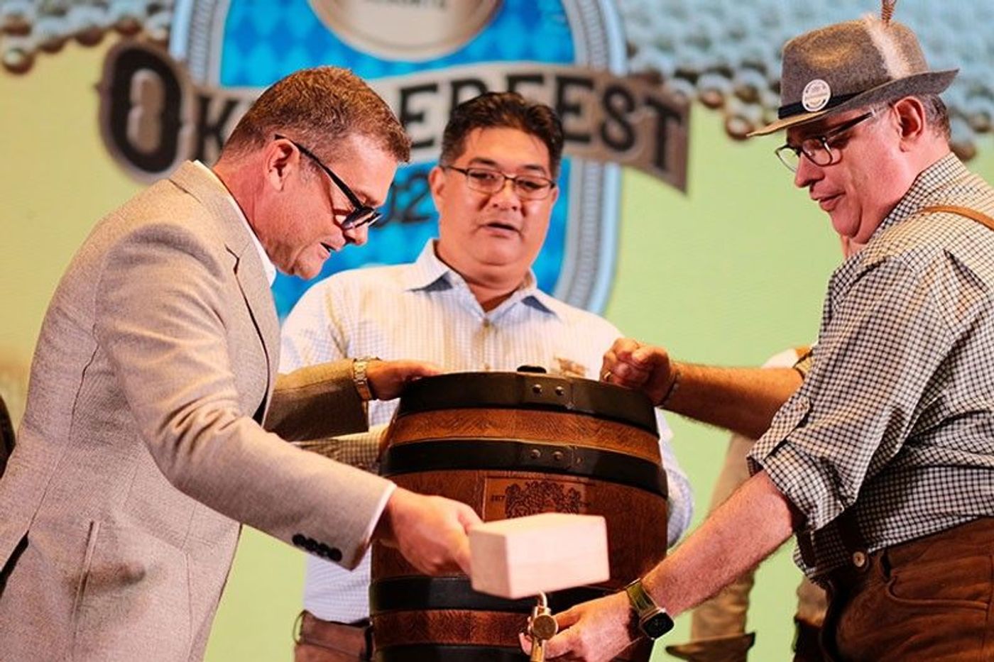 Three men in business and Bavarian attire tap a wooden keg with a mallet on stage beneath an Oktoberfest banner
