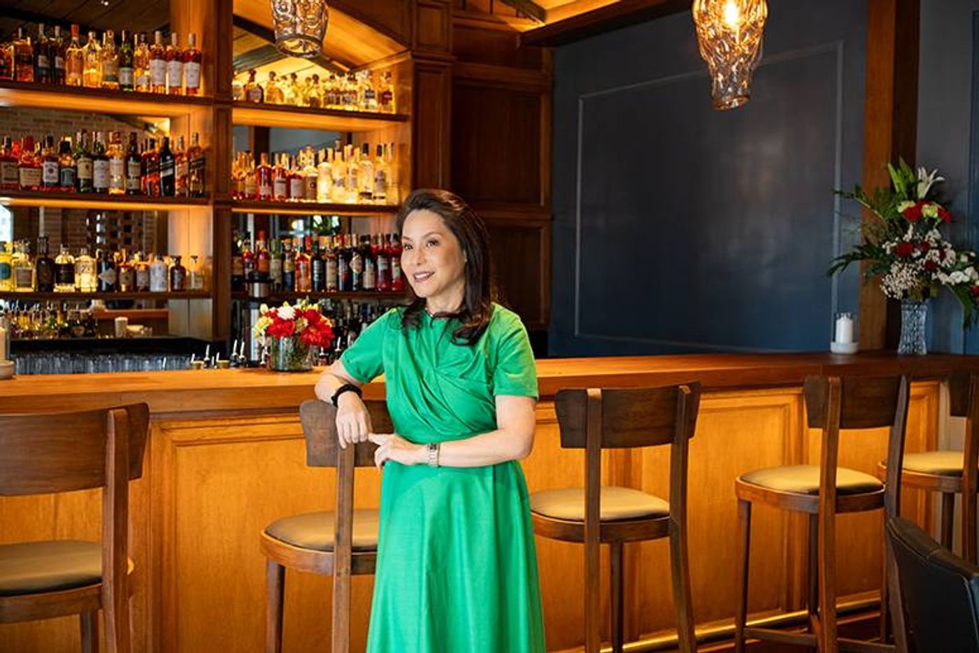 Ana de Ocampo in an emerald dress leans on a chair before Osteria Antica’s amber-lit, bottle-lined bar