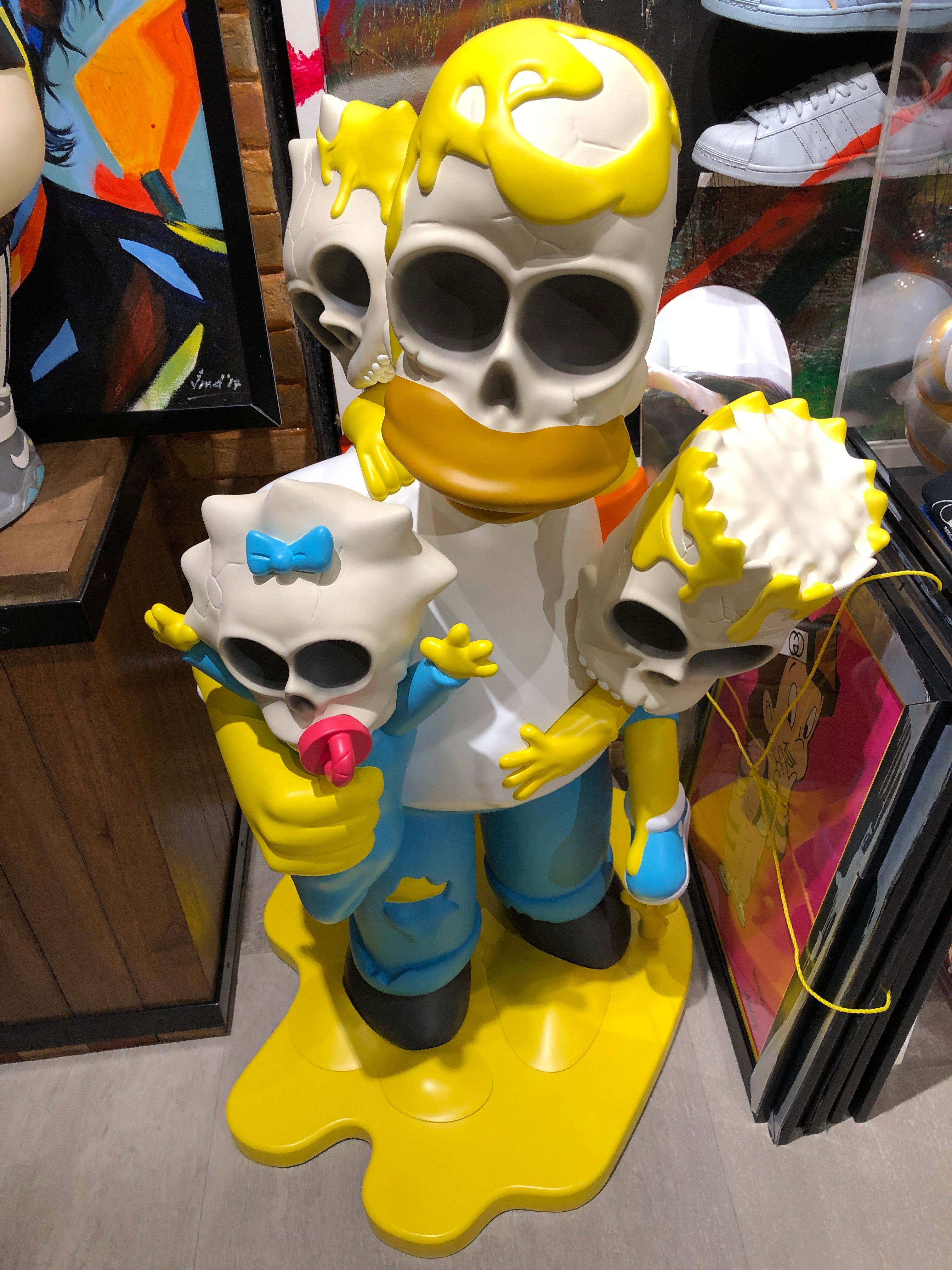 A Simpsons family sculpture with melting yellow skull faces standing in a puddle of yellow, framed art and sneaker boxes on the shelves behind