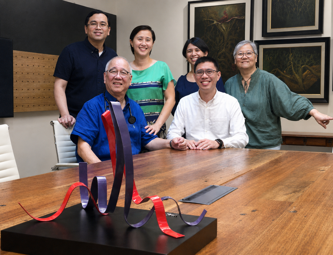 Six members of the Chua family and their designer gathered around a wooden boardroom table, a red and grey ribbon sculpture in the foreground