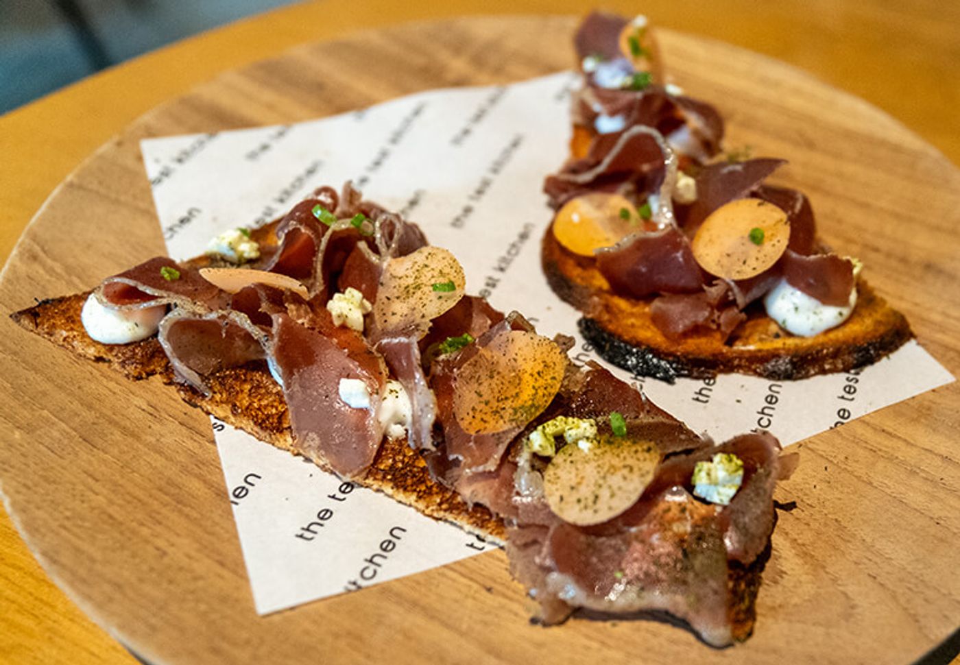 Ribbons of duck prosciutto piled on brioche toasts with dots of garlic emulsion, served on a wooden board