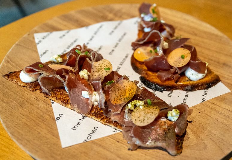 Ribbons of duck prosciutto piled on brioche toasts with dots of garlic emulsion, served on a wooden board