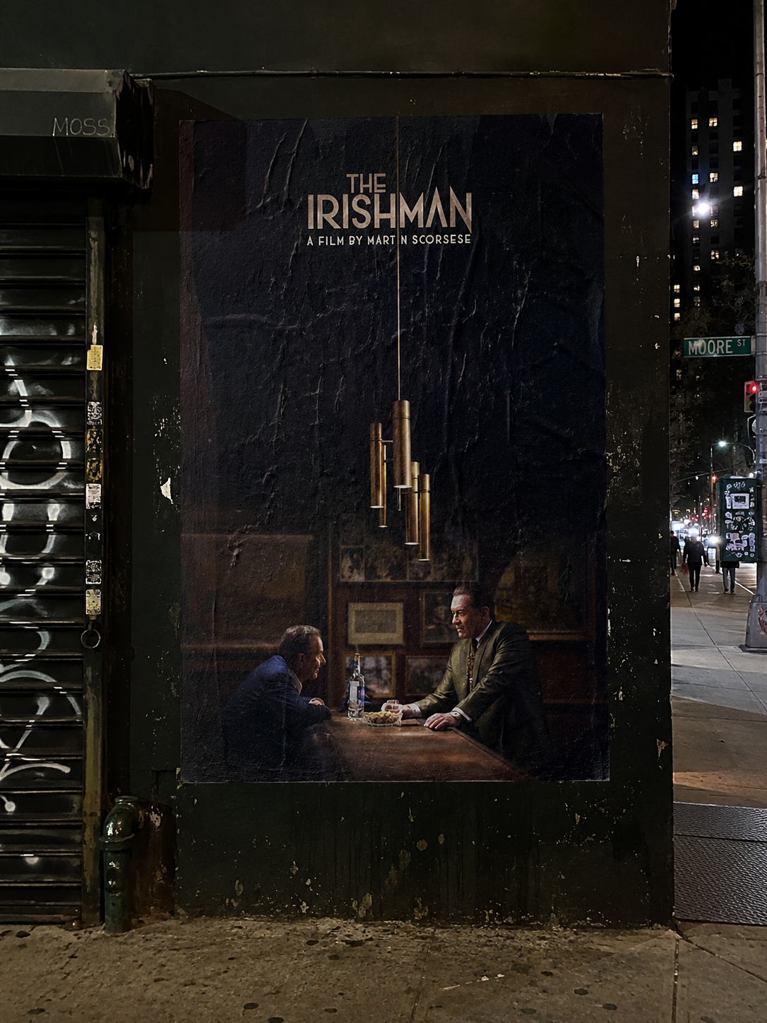 The Irishman movie poster pasted to a dark street-corner wall at night beside a graffiti-covered shutter