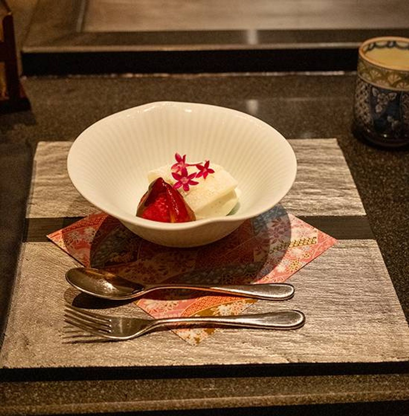 Vanilla ice cream and mochi with red fruit in a fluted white bowl, spoon and fork on a stone slab
