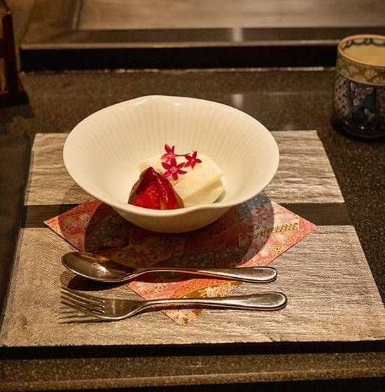 Vanilla ice cream and mochi with red fruit in a fluted white bowl, spoon and fork on a stone slab