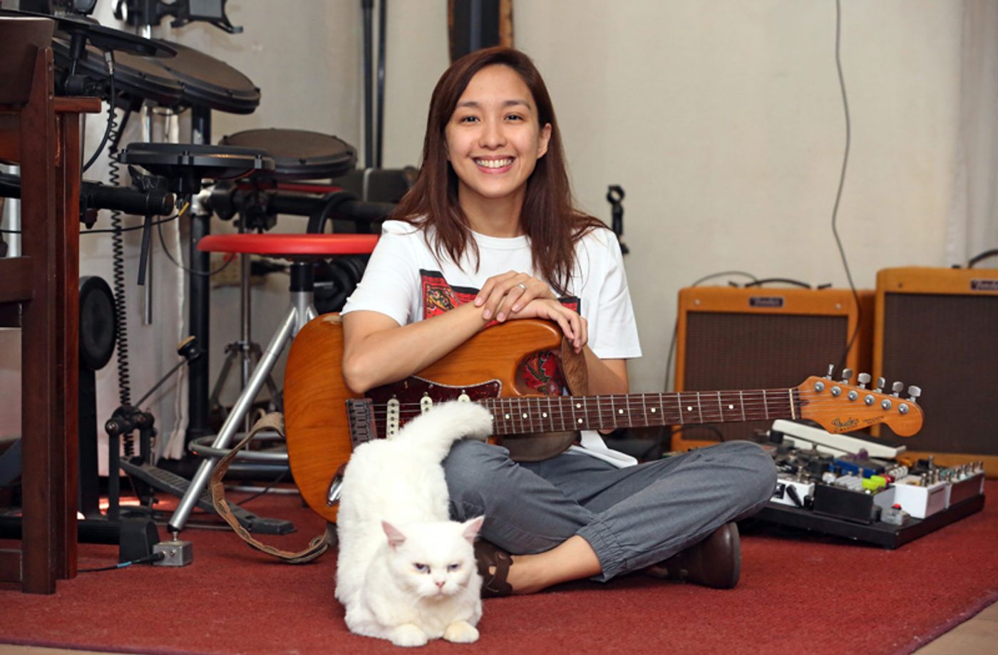 Barbie Almalbis sits cross-legged on a red rug with a sunburst Fender Stratocaster, a white Persian cat stretched out in front of her, a drum kit and amplifiers behind