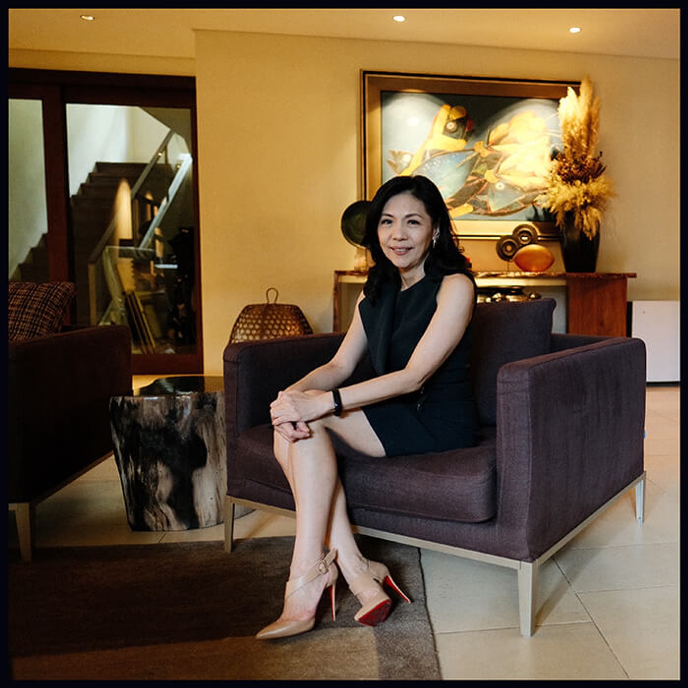 Kaye Tinga sitting in a dark purple armchair in her modern home living room, smiling at the camera with artwork and a console in the background