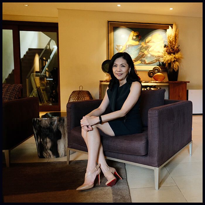Kaye Tinga sitting in a dark purple armchair in her modern home living room, smiling at the camera with artwork and a console in the background