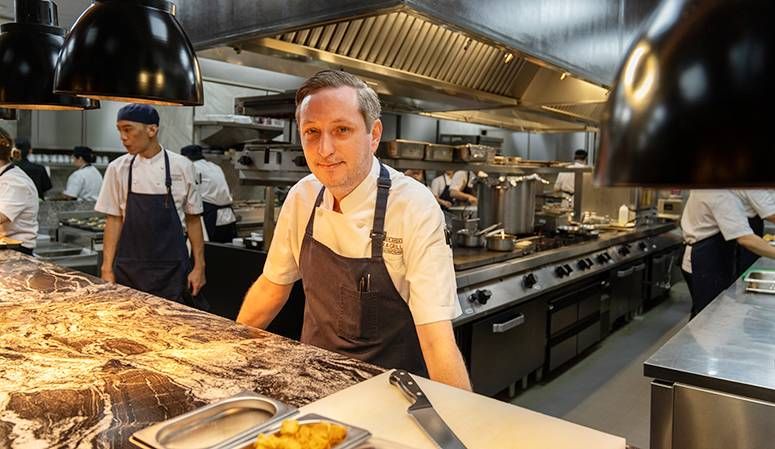 Chef Stephen Wood in an apron at the pass of a gleaming steel kitchen, cooks working behind him