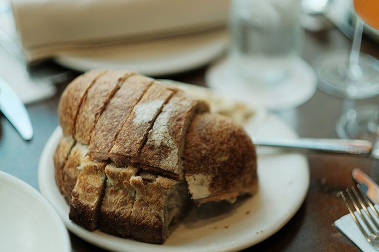 Sliced house-made sourdough loaf on a white plate at a set table with water glasses behind
