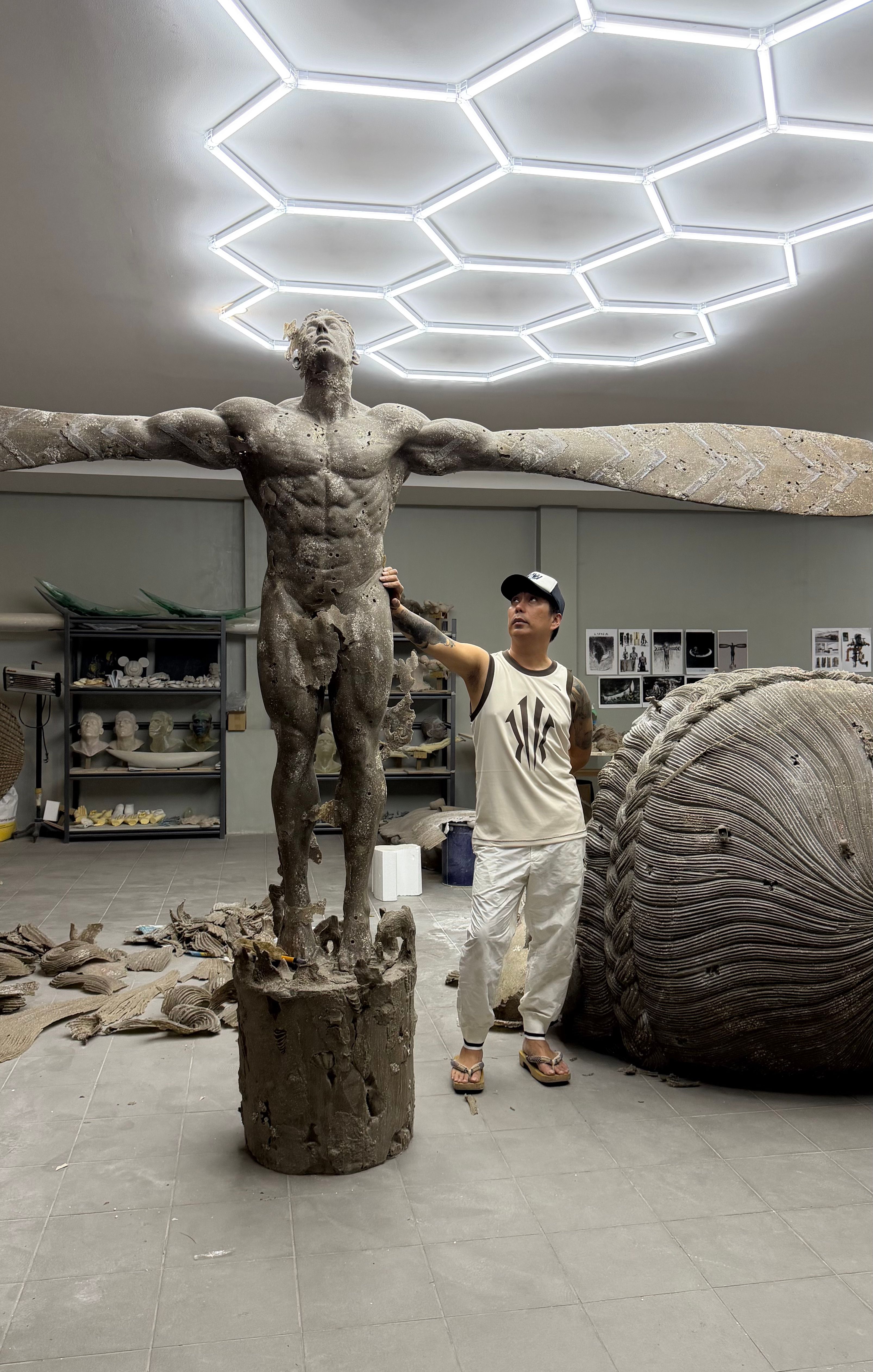 Ronald Ventura in his studio steadying a monumental winged male figure sculpture, a coiled woven form and shelves of casts behind