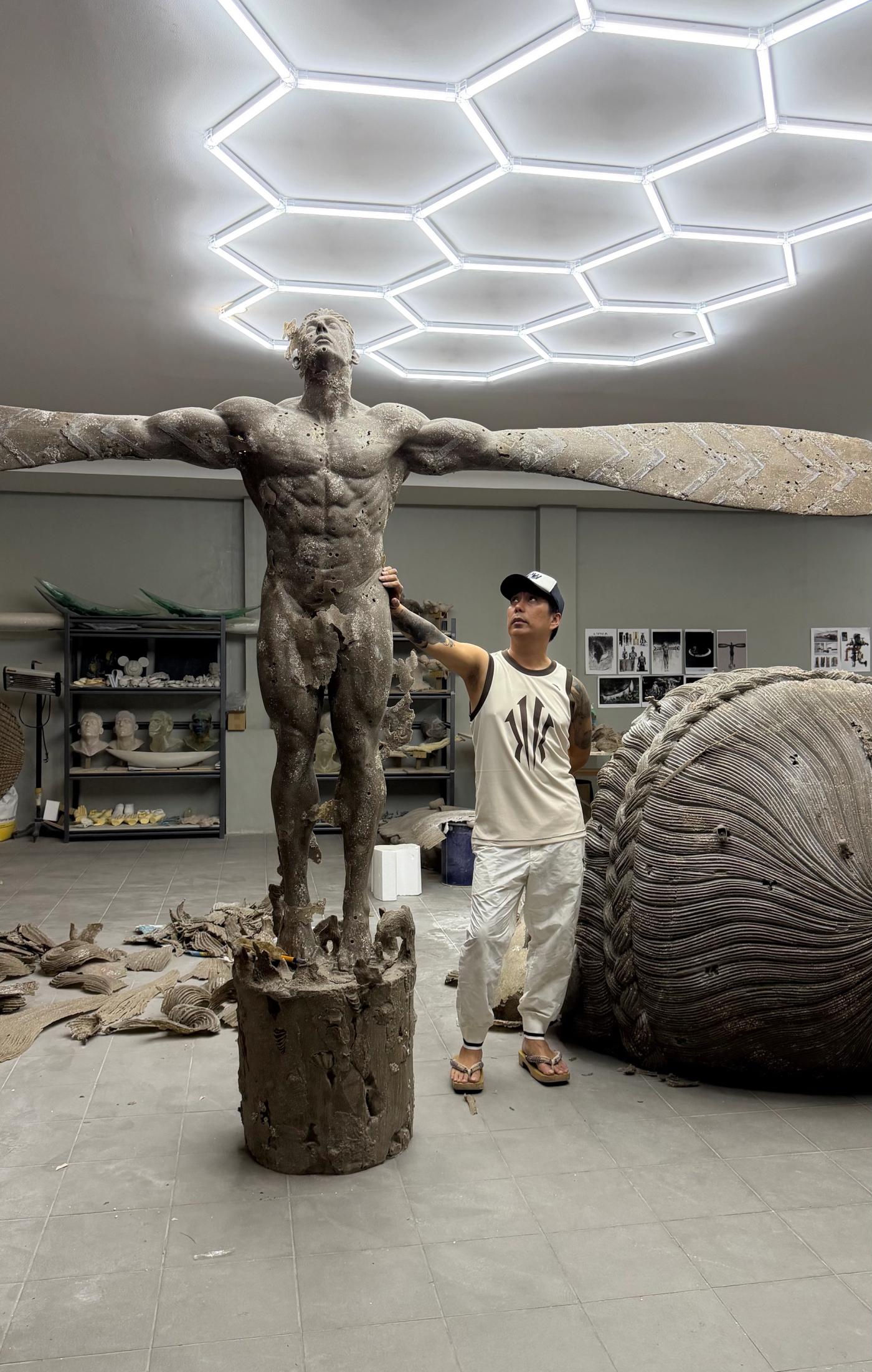 Ronald Ventura in his studio steadying a monumental winged male figure sculpture, a coiled woven form and shelves of casts behind