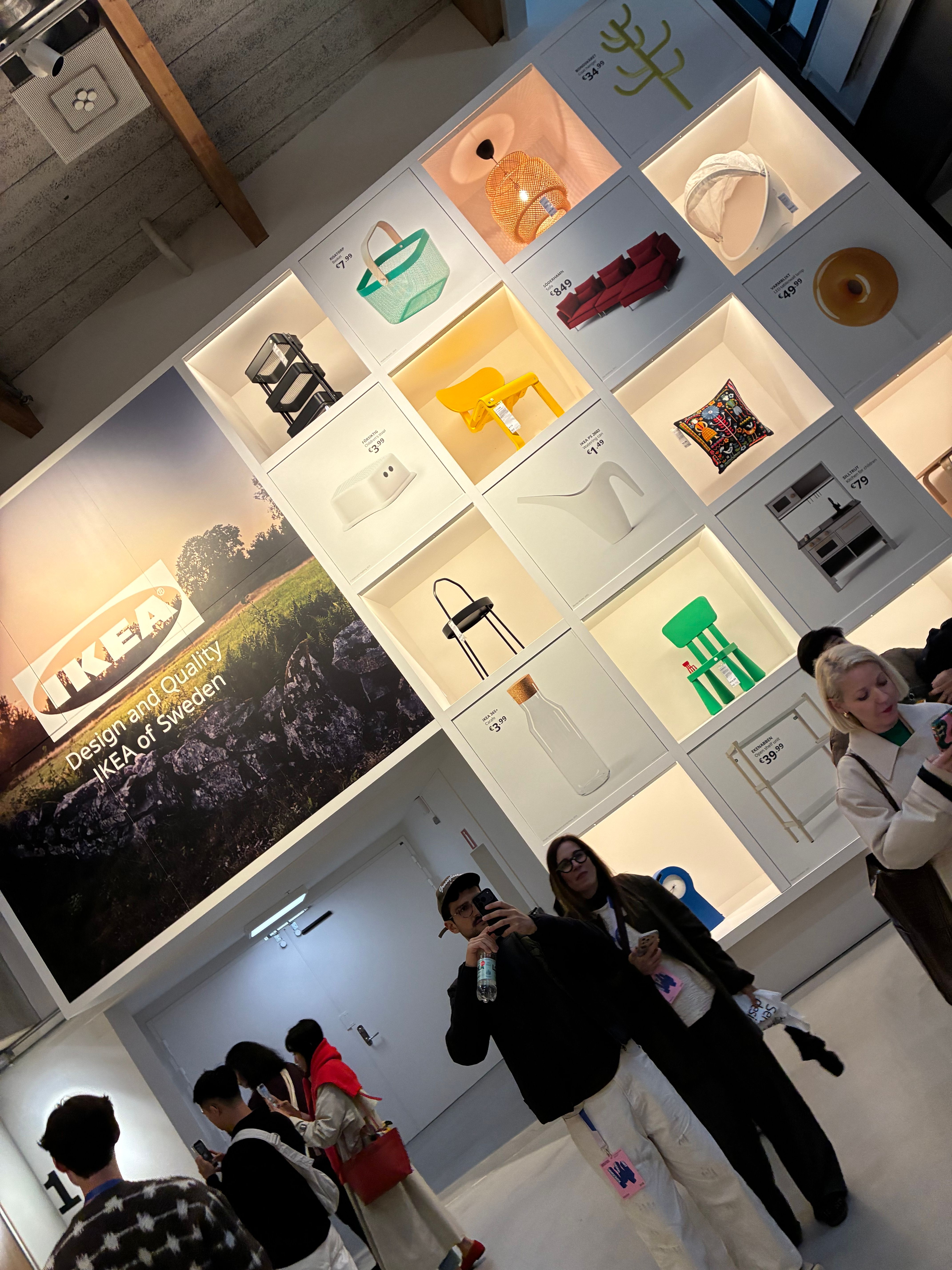 Wall of backlit niches displaying IKEA products beside a Design and Quality IKEA of Sweden mural as visitors browse below
