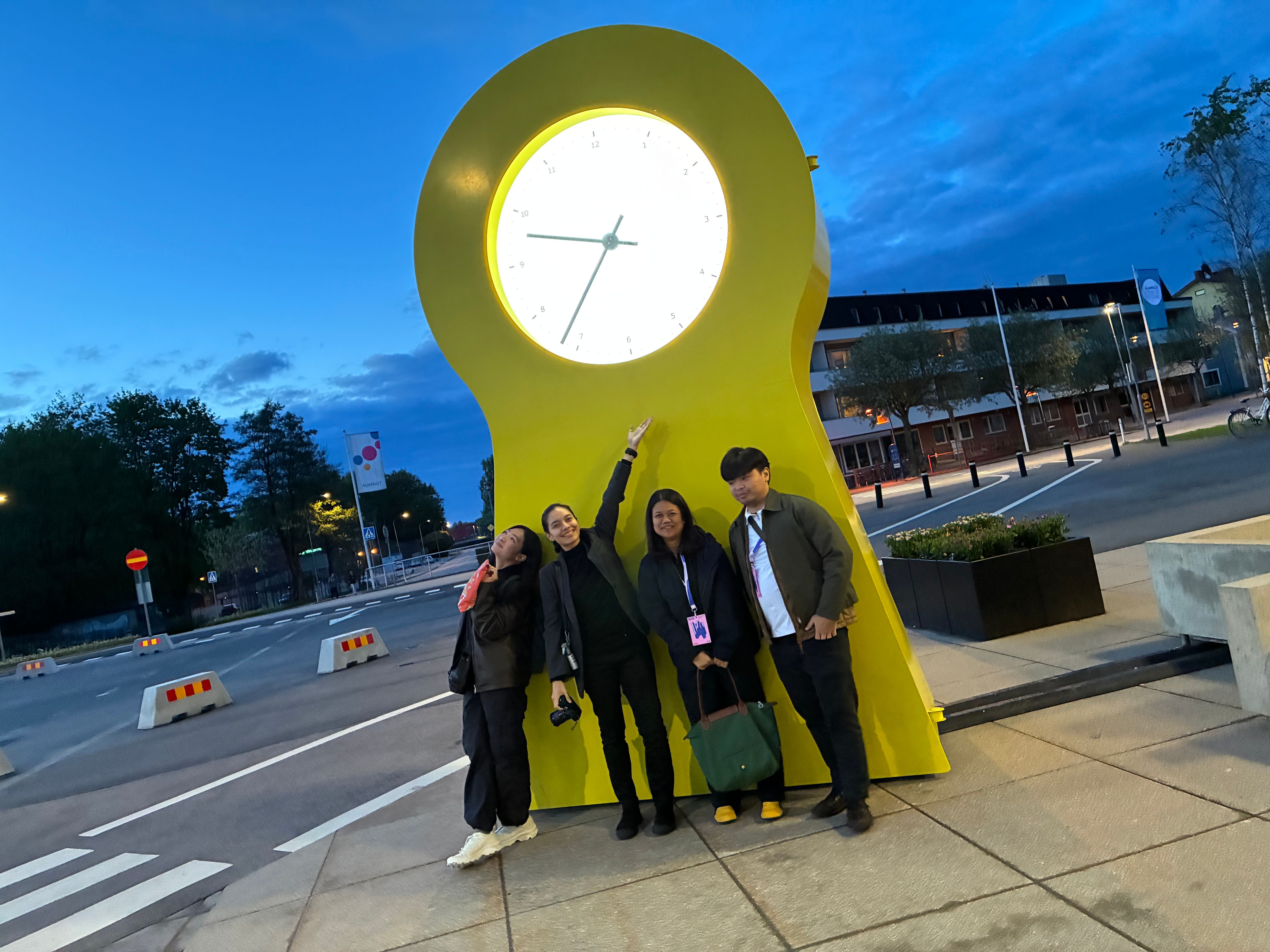 Four visitors posing at dusk with the giant yellow IKEA PS clock sculpture on an Älmhult street corner