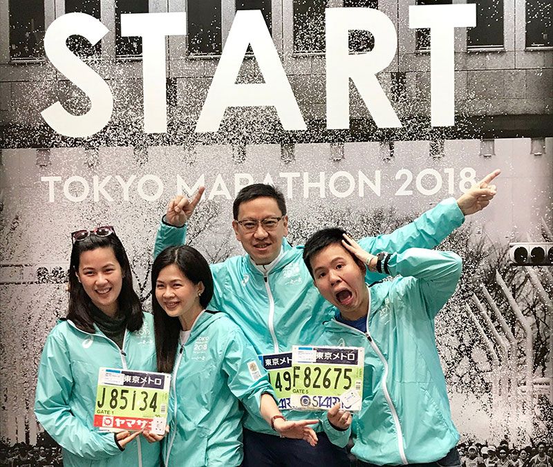 Four runners in matching teal jackets posing with race bibs in front of a large Tokyo Marathon 2018 START backdrop