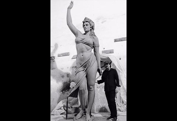 Ginés Serrán in a dark shirt and cap standing beside his towering sculpture Calypso, a draped female figure with one arm raised, in his studio