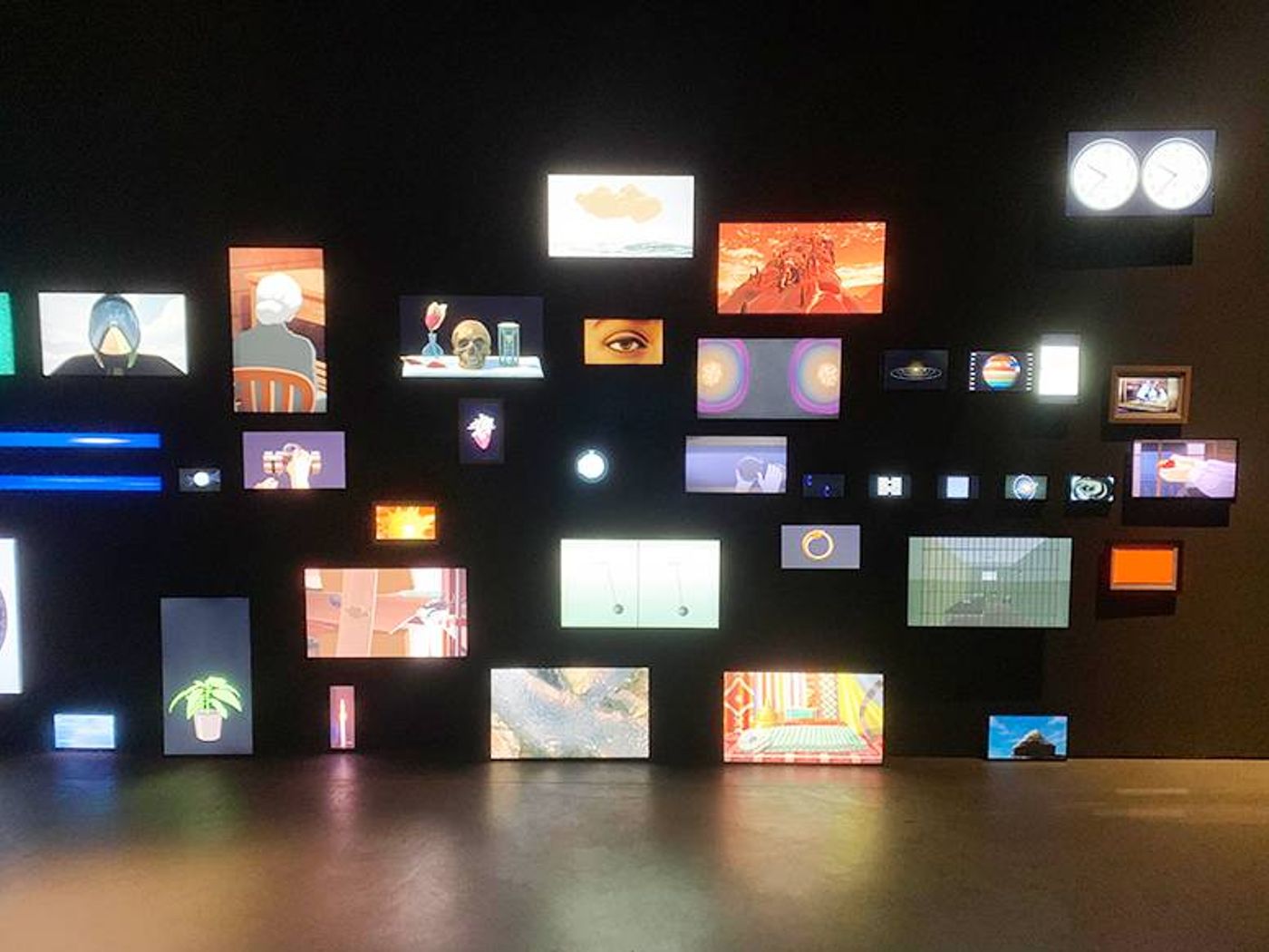 Dozens of small mismatched video screens glowing on a dark gallery wall in Ho Tzu Nyen’s T for Time: Timepieces