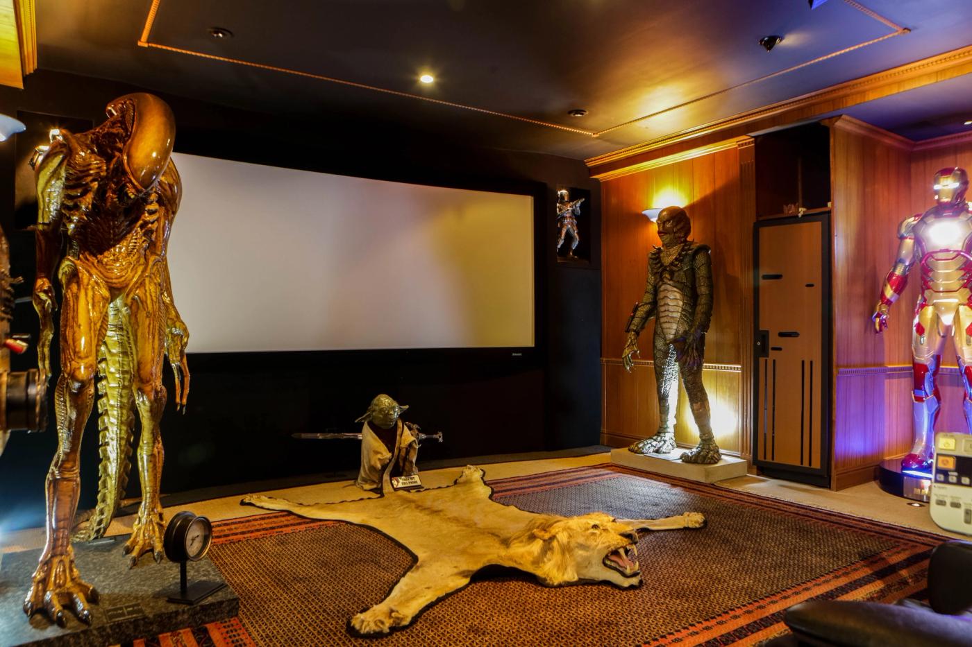 A home cinema with a wide screen, a life-size Alien figure, Yoda, the Creature from the Black Lagoon and an Iron Man suit standing around a lion-skin rug