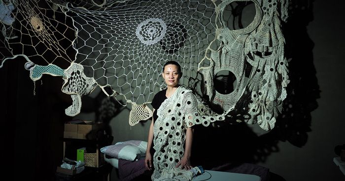 Fiber artist Aze Ong stands beneath her sprawling white crochet installation, webbed panels and rings spanning the dark ceiling, one lacy length draped over her shoulder like a gown