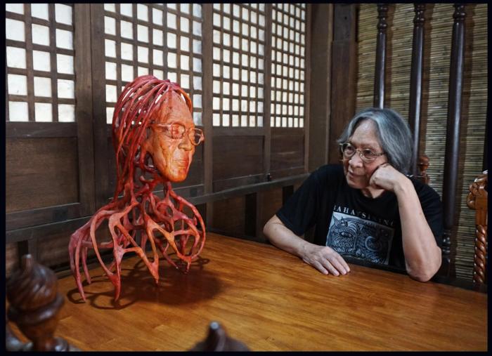 Junyee gazing at his red root-like sculpture of a head on a wooden table by lattice windows