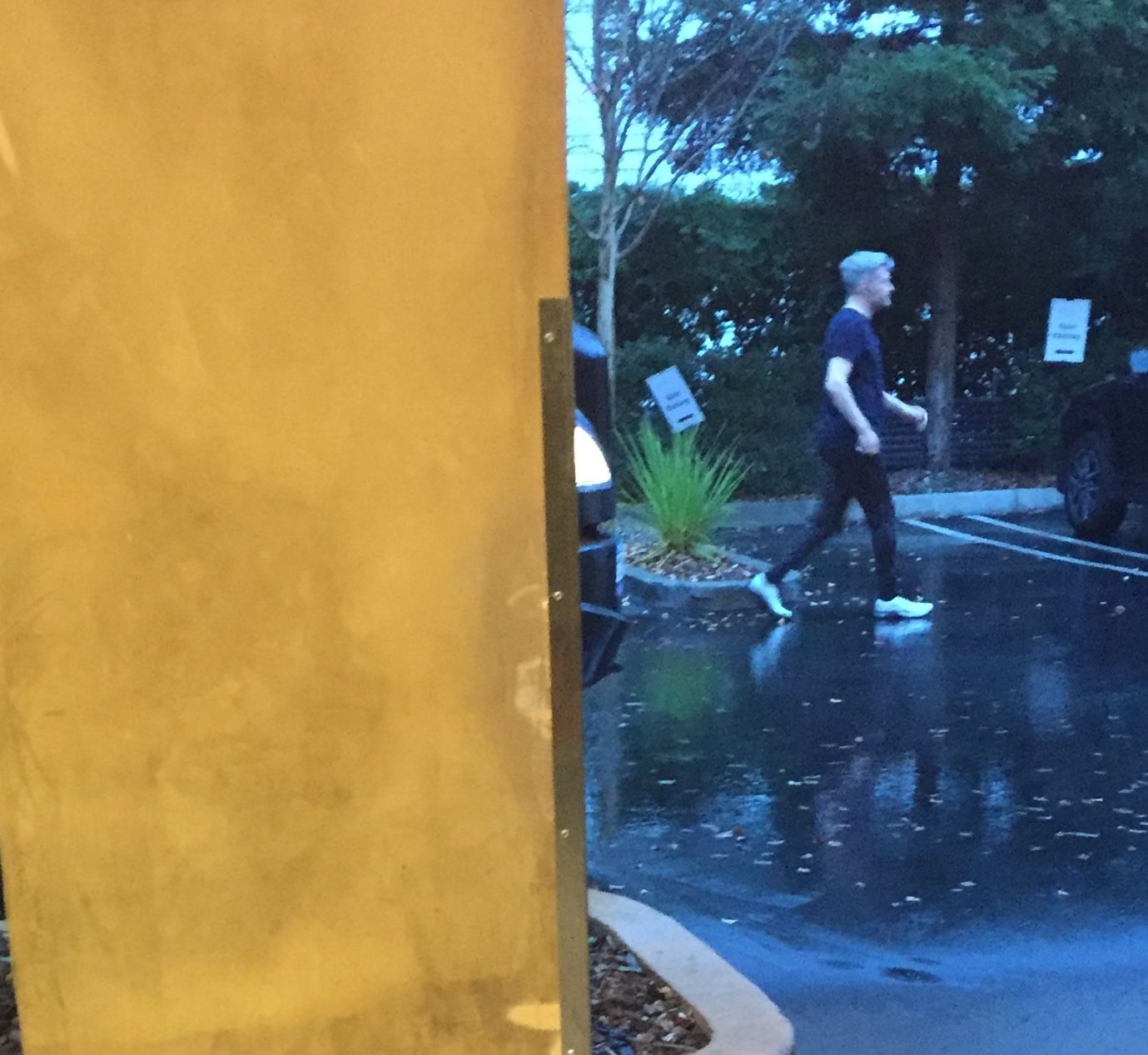A man in dark clothes crosses a rain-slicked parking lot at dusk, glimpsed past a yellow wall